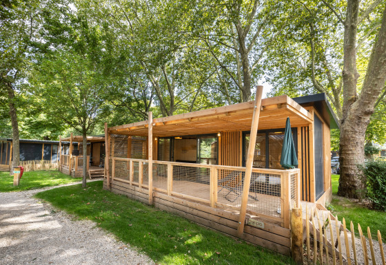 En træbeklædt Mobile Home Vancouver-hytte med overdækket terrasse, omgivet af grønne træer i et ferieområde.