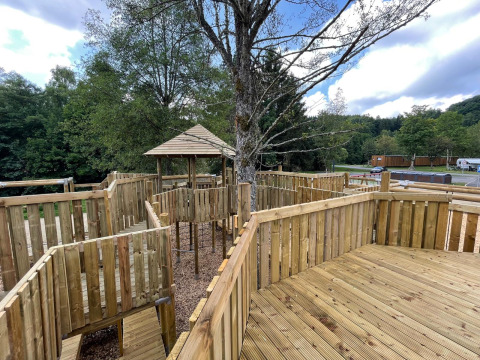 Struttura da gioco in legno con passerelle recintate e gazebo vicino a una tenda safari tra gli alberi.