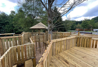 Holzspielplatz mit Überdachung und Geländern in der Nähe eines Safari-Zeltes, umgeben von Bäumen.