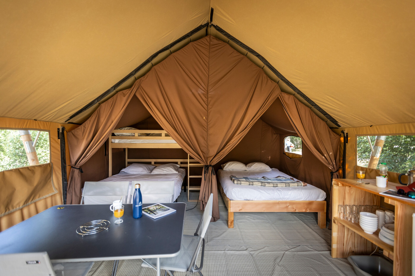 Interieur van een safaritent met tweepersoonsbed, stapelbed, eettafel en planken bij Huttopia Champagny, Frankrijk.