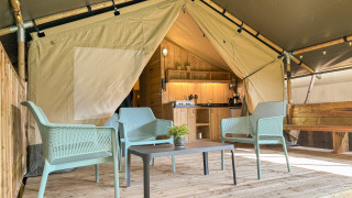 Interior view of a safari tent showing a wooden deck, blue chairs around a table, and a kitchen in the back.