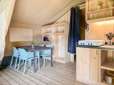Interno tenda safari con tavolo da pranzo, sedie blu, mobili in legno e angolo cottura con fornello.