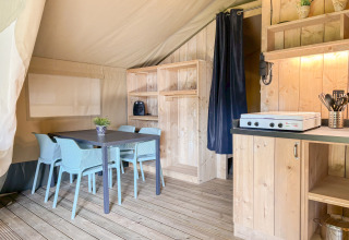 Intérieur de tente safari avec table à manger, chaises bleues, meubles en bois et coin cuisine équipé.
