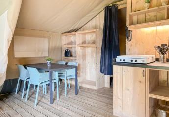 Safari tent interior with dining table, blue chairs, wooden cabinets, and a kitchen area with stove.