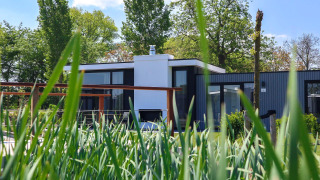 Modern tiny house Cube Exclusive Plus, viewed through tall grass with green trees and blue sky behind.