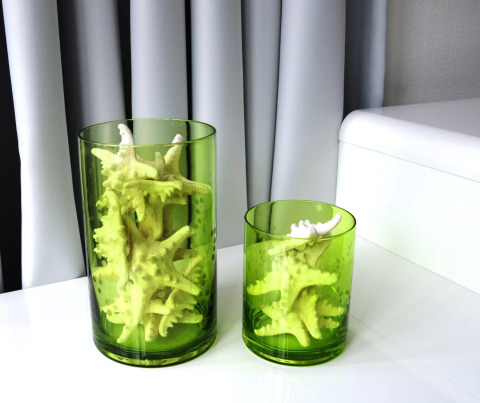 Twee groene glazen vazen met zeesterren erin, op een witte tafel voor lichte gordijnen in het interieur.