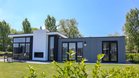 Moderne tiny house Cube Exclusive 4 met grote ramen en terras, omringd door gras en groen op een zonnige dag.