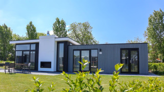 Modern tiny house Cube Exclusive 4 with large windows and terrace, surrounded by greenery on a sunny day.