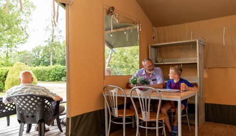 Older man and boy chatting inside a safari tent at De Tien Heugten, Netherlands, with greenery outside.