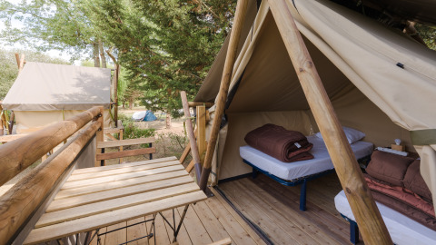 Ein Safarizelt mit zwei Betten und braunen Decken, Holzterrasse mit Tisch und Stühlen im Grünen.