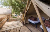 Ein Safarizelt mit zwei Betten und braunen Decken, Holzterrasse mit Tisch und Stühlen im Grünen.
