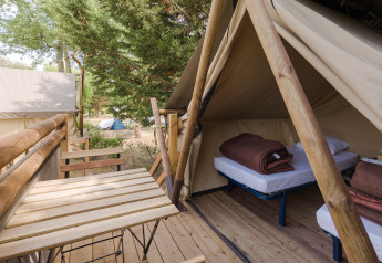 Ein Safarizelt mit zwei Betten und braunen Decken, Holzterrasse mit Tisch und Stühlen im Grünen.