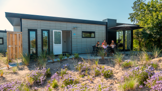 Lodge moderne avec terrasse où des gens se détendent, entourés de fleurs sauvages et de verdure.