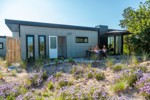 Modern lodge met terras waar mensen buiten zitten, omringd door bloemen en groene natuur.