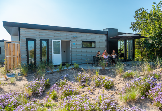 Modern lodge met terras waar mensen buiten zitten, omringd door bloemen en groene natuur.