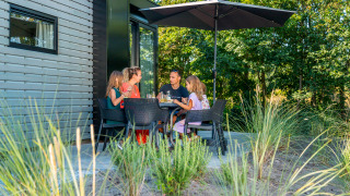 Famille prenant un repas sur la terrasse d'un lodge, protégée par un parasol, entourée de verdure.