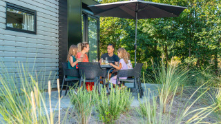 Familia disfruta de una comida al aire libre en la terraza de una cabaña, rodeados de naturaleza.