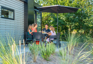 Gezin eet samen op het terras van een lodge onder een parasol, omgeven door groene natuur.