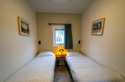 Knusse lodgekamer met twee eenpersoonsbedden, nachtkastje met lamp en een klein raam met gordijn.