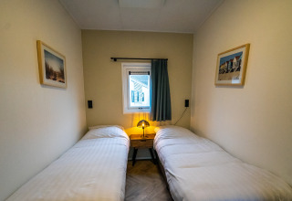 Knusse lodgekamer met twee eenpersoonsbedden, nachtkastje met lamp en een klein raam met gordijn.