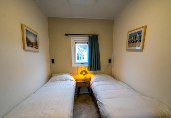 Gezellige lodgekamer met twee aparte bedden, kleine raam, nachtkastje met lamp en wanddecoratie.