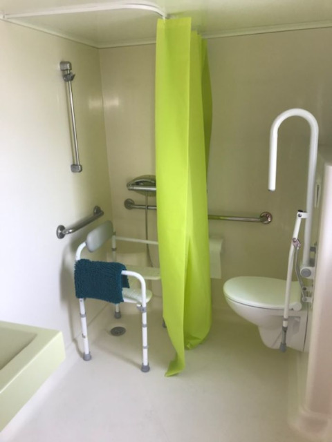 Salle de bain accessible dans un mobil-home avec barres d’appui, siège de douche et rideau vert vif.