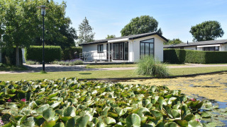 Et moderne sommerhus ved navn Comfort 4 personen MIVA med udsigt over en dam med åkander og grønne træer.