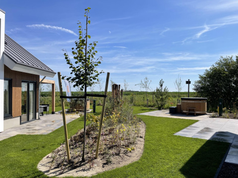 Outdoor area at Duinvilla Wellness lodge with green lawn, young tree, patio, and wooden hot tub on a sunny day.