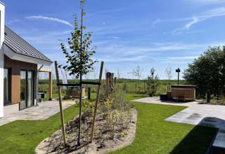 Outdoor area at Duinvilla Wellness lodge with green lawn, young tree, patio, and wooden hot tub on a sunny day.