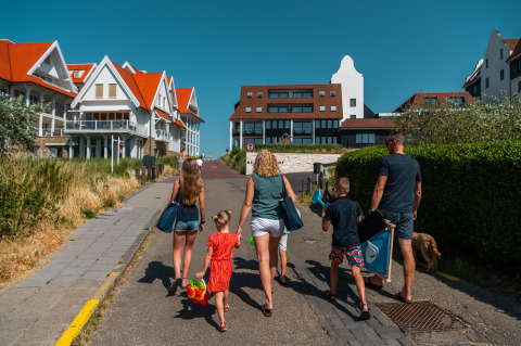 Een gezin wandelt in Cadzand, Zeeland, met strandtassen en speelgoed op een zonnige zomerdag.
