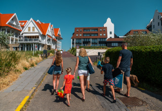 En familie går på en vej i Cadzand, Zeeland, med strandtasker og legetøj, på en solrig dag.