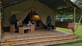 Safari-Zelt Forest Lodge mit Sanitäranlagen, zwei Personen sitzen auf der Terrasse inmitten der Natur.