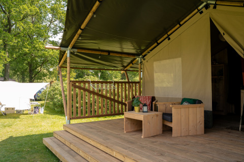 Safari tent veranda met houten zetels, overdekt terras en groene tuin, met sanitaire voorzieningen.