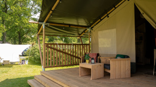 Safari tent porch with wooden furniture, covered deck, and green surroundings, including amenities.