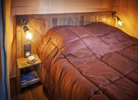 Cozy bed with rustic wood headboard in Lodge Kenya Vintage, Campsite Le Château, France, warm lighting.