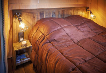 Cozy bed with rustic wood headboard in Lodge Kenya Vintage, Campsite Le Château, France, warm lighting.