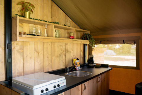 Kleine keukenruimte in een safaritent met houten panelen, kookplaat, gootsteen en open schap met mokken.