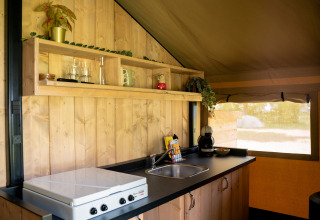 Kleine keukenruimte in een safaritent met houten panelen, kookplaat, gootsteen en open schap met mokken.