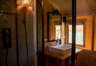 Modernes Badezimmer in einem Safarizelt mit Waschbecken, Spiegel, Dusche und Sonnenlicht am Fenster.