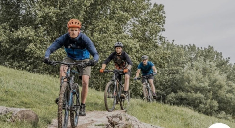 Three cyclists ride mountain bikes on a green trail near Lanaken, Belgium, surrounded by lush trees and nature.