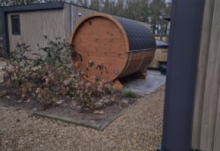Außenfoto einer fassförmigen Sauna neben einer Lodge „Batenburgh“ Deluxe im Veluwe park de Bosgraaf, Niederlande.