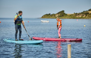 Two people stand on paddleboards in the water at Emmerbølle Strand Camping on a sunny day.