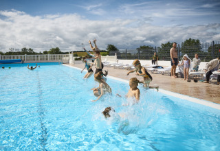 Børn hopper i swimmingpool ved Emmerbølle Strand Camping under en skyet himmel med tilskuere i baggrunden.