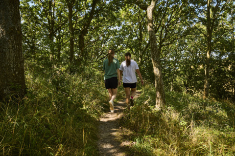 Dos personas caminan por un sendero en el bosque en Emmerbølle Strand Camping, Dinamarca del Sur, rodeadas de árboles.
