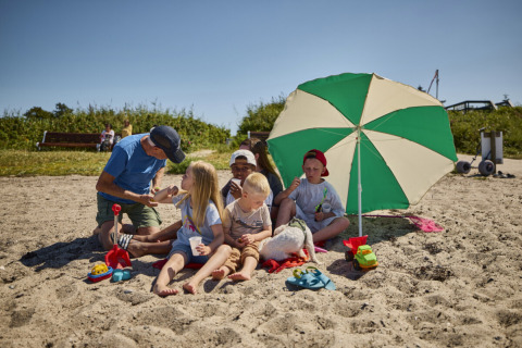 Familie mit Kindern spielt und entspannt unter einem Sonnenschirm am Strand von Emmerbølle Strand Camping.