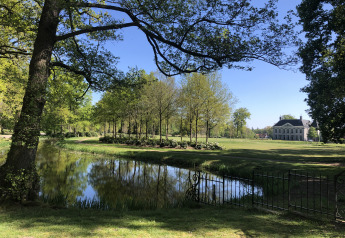 Wunderschöner Glampingplatz mit Teich, grüner Wiese, Bäumen und herrschaftlichem Haus unter blauem Himmel.