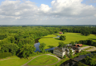 Luchtfoto van een glampingverblijf in een groene omgeving met water, bos en uitgestrekte velden eromheen.