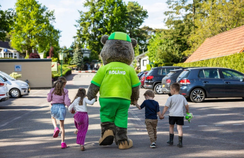 Children walk holding hands with a bear mascot at Campsite Roland in Limburg, Netherlands, among parked cars.