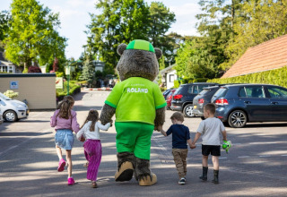 Children walk holding hands with a bear mascot at Campsite Roland in Limburg, Netherlands, among parked cars.