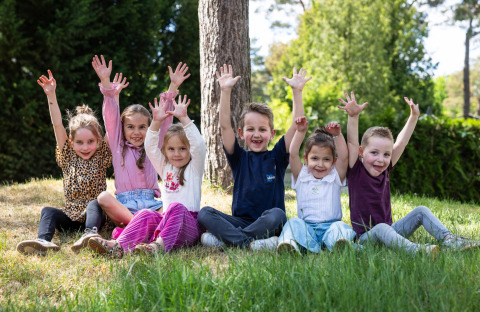 Un gruppo di bambini felici seduti sull’erba con le mani alzate al campeggio Roland a Limburg, Paesi Bassi.